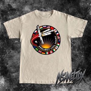 Op de afbeelding: Een beige T-shirt met korte mouwen en een cirkelvormige afbeelding op de borst. Het ontwerp toont een ruimteschip, een heldere zonnestraal en de tekst "PROJECT HAIL MARY" omringd door een rand van diverse internationale vlaggen op een zwarte achtergrond.