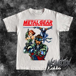 May include: A white short-sleeve t-shirt featuring a graphic print of characters from Metal Gear Solid. The design includes the red text TACTICAL ESPIONAGE ACTION METAL GEAR SOLID above illustrations of a soldier, a woman, and a ninja figure.