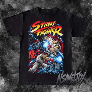 Può includere: Una t-shirt nera con una stampa grafica del personaggio Ryu di Street Fighter. Il design include il testo Street Fighter, il nome Ryu e caratteri kanji giapponesi, circondati da effetti di fulmini blu e scariche di energia.