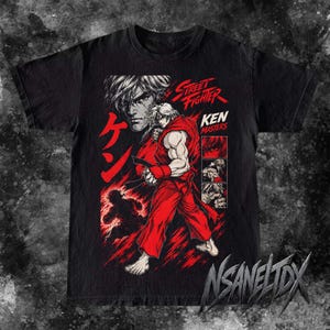 Pode incluir: Uma t-shirt preta de manga curta com uma impressão gráfica da personagem Ken Masters de Street Fighter. O design inclui texto japonês vermelho, as palavras Street Fighter Ken Masters e três painéis inseridos com detalhes da personagem.