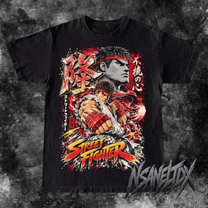 May include: A black short-sleeve t-shirt featuring a graphic print of the character Ryu from Street Fighter. The design includes red and yellow text, Japanese kanji characters, and a dynamic illustration of the fighter in a combat pose.