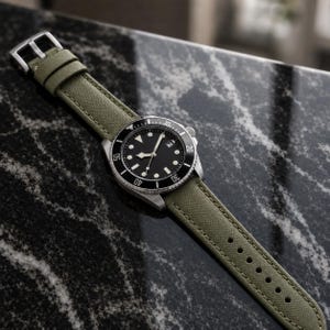 May include: A wristwatch with a black circular dial, luminous hour markers, and a date window at the three o'clock position. The watch features a black rotating bezel with white numerals and is attached to an olive green fabric strap with a silver buckle.