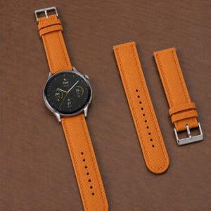May include: A smartwatch with a round black digital display and a bright orange fabric strap, accompanied by two additional matching orange fabric watch strap segments featuring a silver metal buckle and multiple adjustment holes.