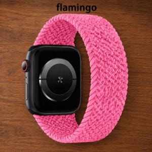 Puede incluir: Un reloj inteligente negro con un sensor circular en la parte posterior y una correa elástica trenzada de color rosa brillante. La palabra flamingo está impresa en negro sobre el reloj. El dispositivo tiene un botón lateral con un anillo rojo.