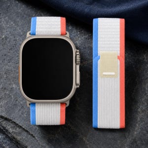 Pode incluir: Um smartwatch com caixa retangular cinza metálico e tela preta, com uma pulseira de nylon trançado. A pulseira tem uma faixa central branca ladeada por bordas azuis e laranjas, com um passador retangular bege no segmento solto da pulseira.