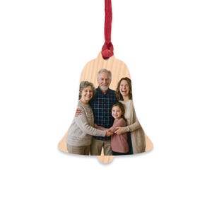 Può includere: Un ornamento in legno a forma di campana con una fotografia stampata di una famiglia di quattro persone. L'ornamento ha una finitura in venature di legno chiaro ed è sospeso da un sottile nastro rosso attaccato alla parte superiore. Il gruppo familiare è composto da due adulti e due bambini.