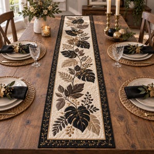 May include: A long rectangular table runner featuring a cream-colored center panel with a quilted pattern of dark brown and gold monstera leaves and foliage. The runner is bordered by a black fabric edge with gold embroidered vine details.