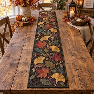 May include: A rectangular fabric table runner featuring a dark background with a detailed quilted pattern of colorful autumn leaves, including red maple, yellow ginkgo, and green oak leaves, interspersed with small brown acorns and berries.