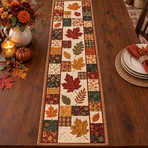Può includere: Un runner da tavola trapuntato rettangolare con un pannello centrale in tessuto color crema decorato con varie foglie autunnali applicate nei toni del rosso, arancione e verde, bordato da un patchwork di quadrati con motivi floreali e scozzesi.