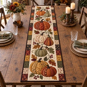 May include: A rectangular fabric table runner featuring a quilted design with various patterned pumpkins, autumn leaves, and berries in shades of orange, green, and cream. The border consists of a patchwork pattern of small squares in autumnal colours.