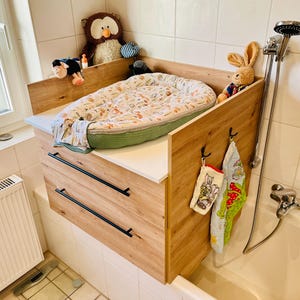May include: A light wood changing table topper with two drawers, featuring a white top surface and a patterned baby nest. The unit includes black handles, side panels with hooks holding two small towels, and several plush toys placed on top.