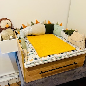 May include: A wooden changing table with a drawer, featuring a white side storage box containing a brown owl plush toy. A long, multi-colored dinosaur-shaped bumper surrounds a white mattress pad with animal prints and a solid yellow rectangular mat.
