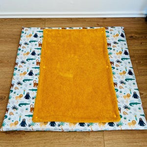 May include: A rectangular play mat featuring a white base with a repeating pattern of lions, monkeys, and crocodiles in shades of orange, brown, and green. A solid mustard yellow textured fabric panel is centered on top of the patterned mat.
