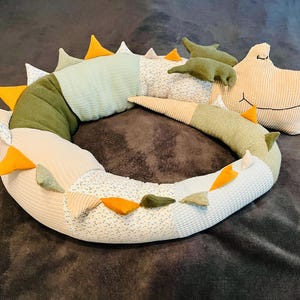 Puede incluir: Un cojín largo y curvado con forma de dragón que presenta un diseño de patchwork en telas de color verde salvia, crema y estampado floral. Tiene una cabeza color crema con rasgos faciales bordados, pequeñas alas verdes y una serie de púas triangulares en naranja, verde y blanco a lo largo de su espalda.