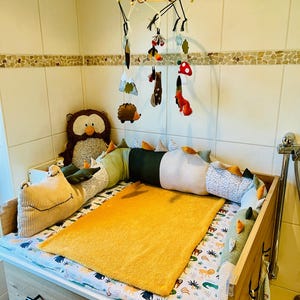 May include: A wooden changing table with a patterned mattress pad featuring animal illustrations, a solid mustard yellow towel, and a plush owl. A fabric mobile hangs above with felt forest elements including a hedgehog, fox, and mushroom.