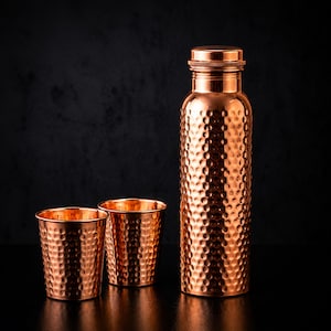 Op de afbeelding: Een hoge, cilindrische koperen waterfles met een gehamerde textuur en een bijpassende schroefdop, vergezeld door twee kleinere, bijpassende gehamerde koperen bekers. De set heeft een gepolijste, metalen afwerking met een opvallend kuiltjesoppervlak.