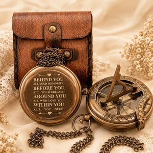 Puede incluir: Una brújula redonda de latón con cadena y estuche de cuero marrón. La tapa de la brújula tiene grabado el texto: BEHIND YOU ALL YOUR MEMORIES BEFORE YOU ALL YOUR DREAMS AROUND YOU ALL WHO LOVE YOU WITHIN YOU ALL YOU NEED. Se muestra una segunda brújula abierta.