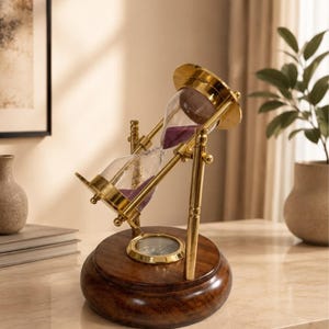 May include: A rotating hourglass featuring a polished gold-tone metal frame and a circular dark brown wooden base. The glass bulb contains purple sand, and the frame is supported by vertical metal pillars, allowing the hourglass to tilt and rotate.