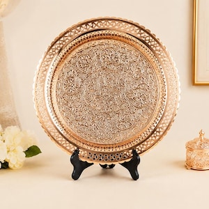 Pode incluir: Uma bandeja decorativa redonda em tom dourado metálico com um intrincado padrão floral e de vinhas em relevo na superfície. A bandeja tem uma borda larga, recortada e rendilhada, e é exibida na vertical sobre um suporte preto.