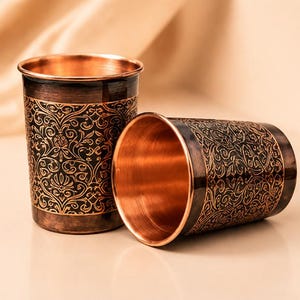 May include: Two cylindrical drinking cups made of polished copper. One cup stands upright while the other rests on its side. Both feature a dark, weathered exterior finish with an intricate, repeating floral pattern etched in bright copper.