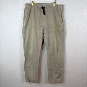 May include: Beige pants featuring an elastic waistband with a black drawstring closure. These trousers have a straight-leg cut, side pockets, and visible stitching details along the seams. The fabric appears to be a cotton-based twill material.
