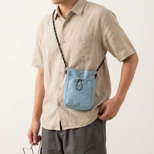 May include: A light blue crossbody bag with a drawstring closure and a black and white patterned cord strap. The bag features a rectangular shape with a front pocket and a small metal snap button at the top center, worn over a beige button-down shirt.