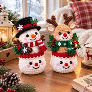 May include: Two handmade crochet amigurumi figures featuring a snowman wearing a black top hat and a reindeer with brown antlers. Both figures are decorated with green foliage, red berries, and white snowflakes, sitting on a wooden surface.