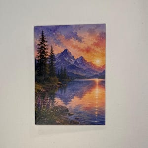 May include: A rectangular print featuring a watercolor landscape of a mountain range reflected in a calm lake at sunset. The sky displays orange, pink, and purple hues. Tall evergreen trees stand on the left shore, with purple wildflowers in the foreground.