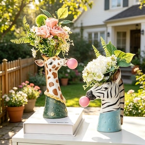 May include: Two ceramic animal-shaped vases, one a giraffe with brown spots and a green base, the other a zebra with black stripes and a blue base. Both figures feature a pink bubble in their mouths and hold bouquets of artificial flowers.