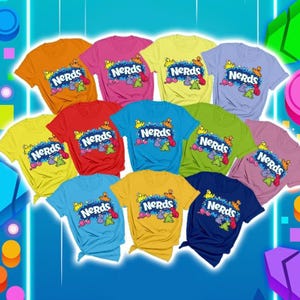May include: A collection of eleven short-sleeved t-shirts in various solid colours, each featuring the word "NERDS" printed in white letters on a blue background, surrounded by small, colourful cartoon character illustrations.