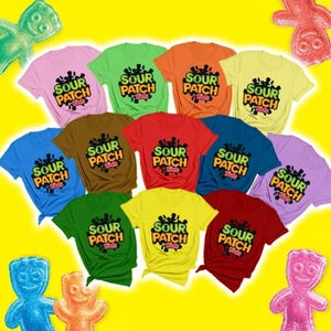 Può includere: Una collezione di undici magliette a maniche corte in vari colori a tinta unita, ognuna con il logo Sour Patch Kids nero e verde sul petto, disposte su uno sfondo giallo brillante con accenti a forma di personaggio di caramella.
