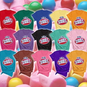 May include: A collection of fifteen short-sleeved t-shirts in various solid colors, each featuring the Dubble Bubble logo printed on the chest. The logo consists of a red oval shape with the text Dubble Bubble and a crown icon above it.