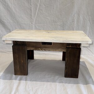 May include: A small rectangular wooden bench featuring a distressed white painted top surface and four thick, dark brown stained wooden legs. The construction shows visible wood grain texture throughout the piece, with a dark metal bracket visible on the front apron.