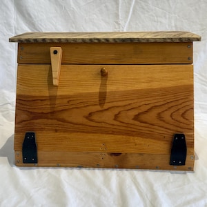 Può includere: Un baule rettangolare in legno con finitura a venature color marrone chiaro. Il baule presenta un coperchio incernierato, un piccolo pomello in legno, una chiusura in legno chiaro sul lato sinistro e due cerniere in metallo nero sul davanti.