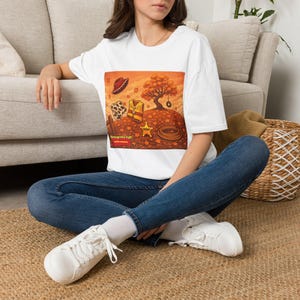 Peut inclure: Un t-shirt blanc à manches courtes avec une impression graphique représentant un paysage d'automne, un chapeau de cowboy marron, un gilet à motif vache, une étoile de shérif jaune et un lasso. Le texte "Through the fall and the summer" est présent.
