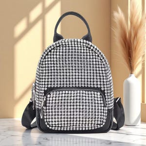 Könnte beinhalten: Ein kleiner Rucksack, der mit einem Gitter aus funkelnden silbernen Strasssteinen bedeckt ist. Die Tasche verfügt über einen schwarzen Tragegriff, schwarze verstellbare Schultergurte und eine Fronttasche mit Reißverschluss. Boden und Besatz bestehen aus glattem schwarzem Material mit silbernen Reißverschlüssen.