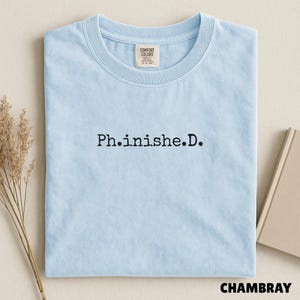 May include: A light blue short-sleeve t-shirt with a crew neckline. The center of the chest features the black embroidered text Ph.inishe.D. The interior neck label displays the brand name Comfort Colors and the size S-P-CH.