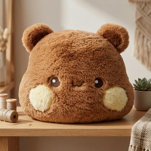 May include: A plush brown bear-shaped pillow with soft, fuzzy fur, two rounded ears, and a small embroidered face. The pillow features two prominent, circular light yellow patches on the cheeks, giving it a distinct, rounded appearance.