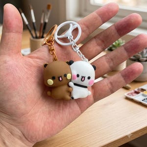 May include: Two cartoon bear keychains, one brown and one white, each featuring a small round face with pink cheeks and a wide open mouth. The brown bear is attached to a brown ring, while the white bear is attached to a white ring, both connected by chains.