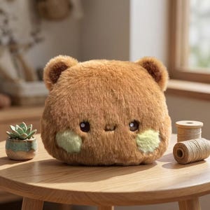 May include: A round, brown plush toy shaped like a bear with two small ears, dark eyes, and light green circular patches on its cheeks. The plush has a soft, fuzzy texture and a simple, neutral expression, sitting on a wooden surface.