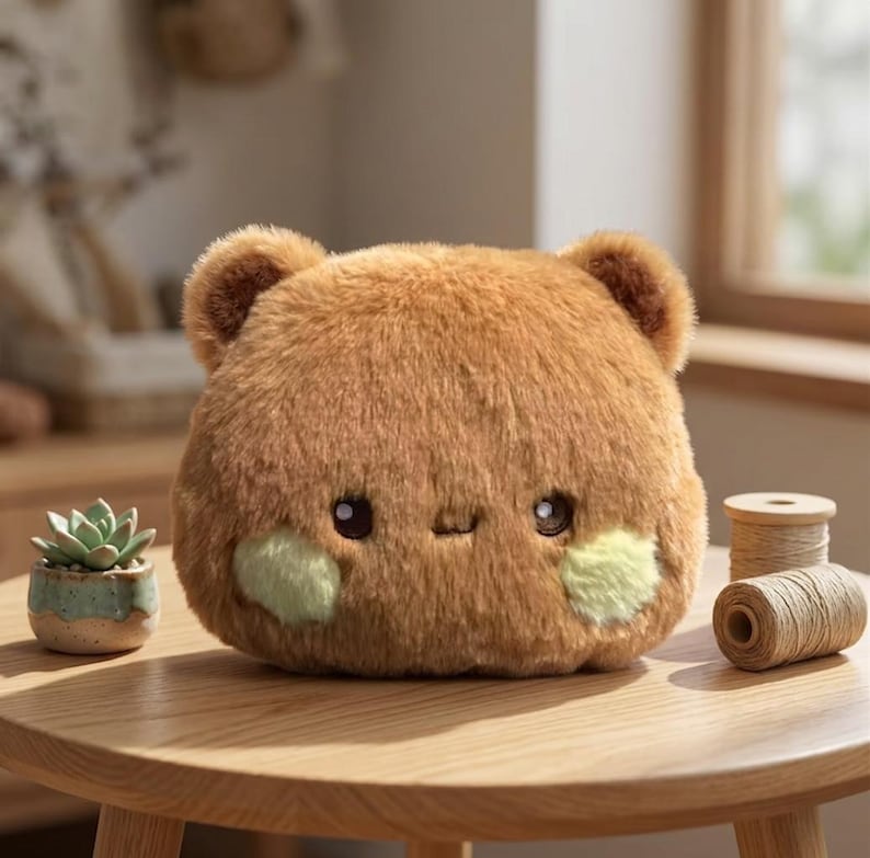 May include: A round, brown plush toy shaped like a bear with two small ears, dark eyes, and light green circular patches on its cheeks. The plush has a soft, fuzzy texture and a simple, neutral expression, sitting on a wooden surface.