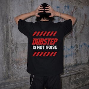 May include: A black short-sleeved t-shirt featuring a graphic on the back. The design includes red diagonal hazard stripes at the top and bottom, with the red text DUBSTEP and the white text IS NOT NOISE centred between the stripes.
