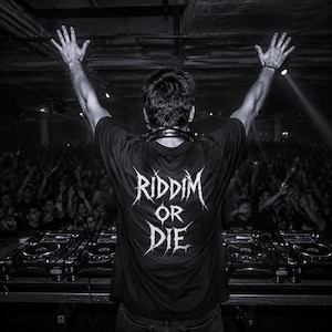 Op de afbeelding: Een zwart t-shirt met de witte tekst "RIDDIM OR DIE" in een gestileerd, grillig lettertype op de achterkant. Het shirt wordt gedragen door een persoon die achter een DJ-mengpaneel met meerdere decks staat en naar een menigte kijkt met armen omhoog.