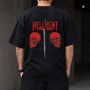 Può includere: Una t-shirt nera a maniche corte con una grafica sul retro. Il design include il testo rosso HELLBENT sopra una spada argentata centrale, affiancata da due teschi rossi. Una linea orizzontale di filo spinato corre dietro la spada e i teschi.