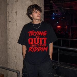 May include: A black short-sleeved t-shirt featuring bold red text on the front that reads TRYING TO QUIT THIS DAMN RIDDIM in a stylised font. The shirt has a crew neckline and is worn with a silver chain necklace and black cargo trousers.