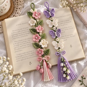 Puede incluir: Dos marcapáginas de ganchillo hechos a mano sobre un libro abierto. Uno presenta flores rosas y blancas con hojas verdes y una borla rosa y blanca. El otro muestra mariposas moradas y blancas con pequeñas cuentas blancas y una borla morada.