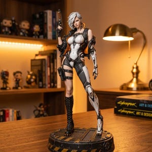 以下が含まれることがあります： 短い白髪のキャラクターの精巧なコレクターズフィギュア。白と黒のボディスーツ、オレンジ色の裏地が付いた黒いジャケット、背の高い黒いブーツを着用しています。キャラクターはハンドガンを持ち、円形の工業用ベースの上に立っています。