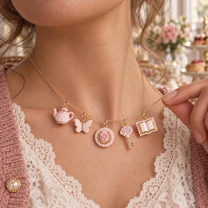 Peut inclure: Un collier à chaîne dorée avec cinq breloques pendantes : une théière rose, un papillon rose, un emblème circulaire à rose rose, une clé ornée rose et un livre ouvert rose. Les breloques présentent des détails délicats blancs et dorés.