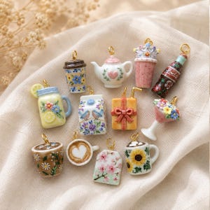 Può includere: Una collezione di ciondoli in ceramica in miniatura con vari design, tra cui una teiera, tazze da caffè con motivi floreali, un cartone del latte con uccellini, una bottiglia di soda, un regalo, un barattolo di limone e un bouquet, con anelli dorati.