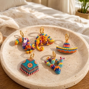 Puede incluir: Una colección de cinco dijes de metal coloridos que representan un tucán, un camaleón, un sombrero, un poncho y un cactus. Cada dije tiene acabados con patrones de esmalte vibrantes e incluye una argolla dorada para sujetar a joyas.